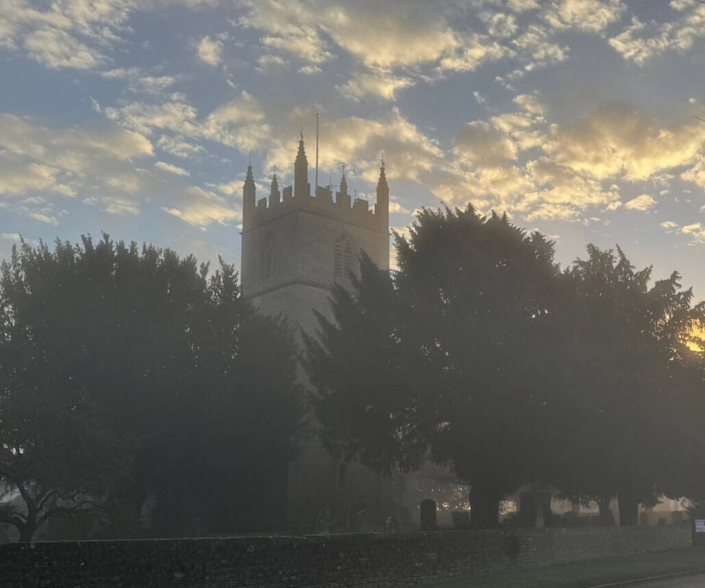 A church tower rises above trees at sunrise, with mist and clouds in the sky.